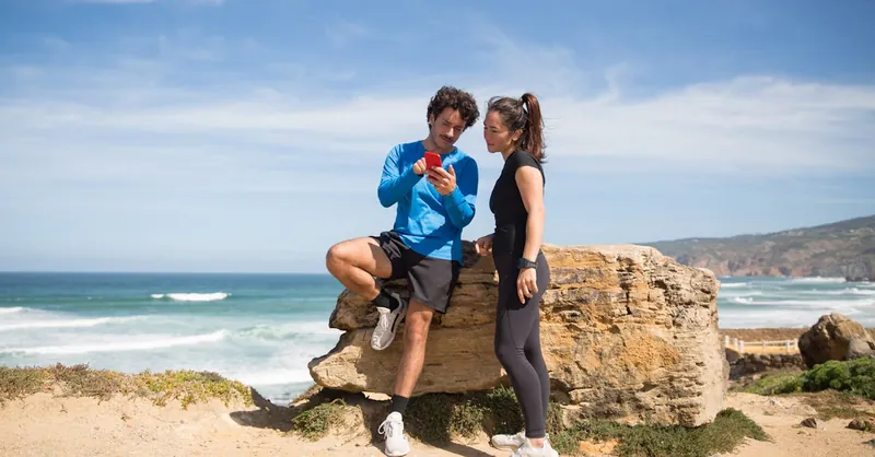 Fit couple taking a break on rocky beach in Portugal, using smartphone for fitness tracking.