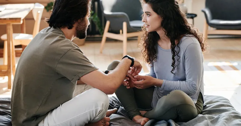 A couple sitting closely on a bed, engaged in a thoughtful discussion in a cozy home setting.