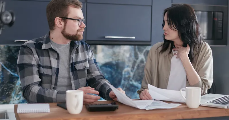 A young couple sitting at a table discussing bills and financial plans in a modern kitchen.