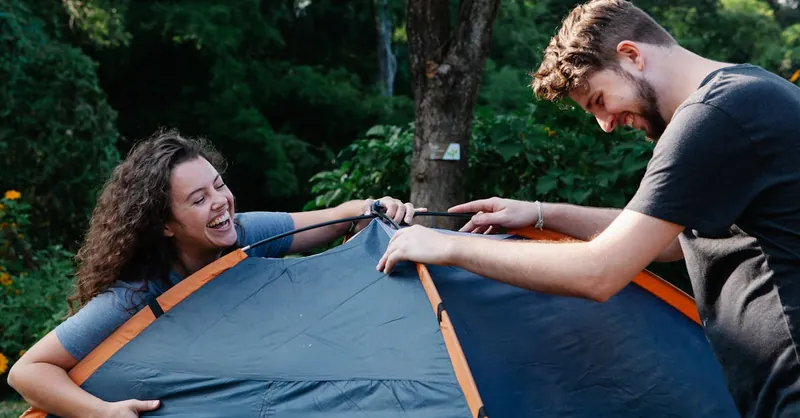 Cheerful young couple in casual clothes laughing while setting up camping tent during romantic picnic in nature
