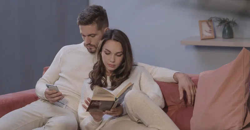 A couple enjoying leisure time with a book and smartphone on a comfortable sofa at home.