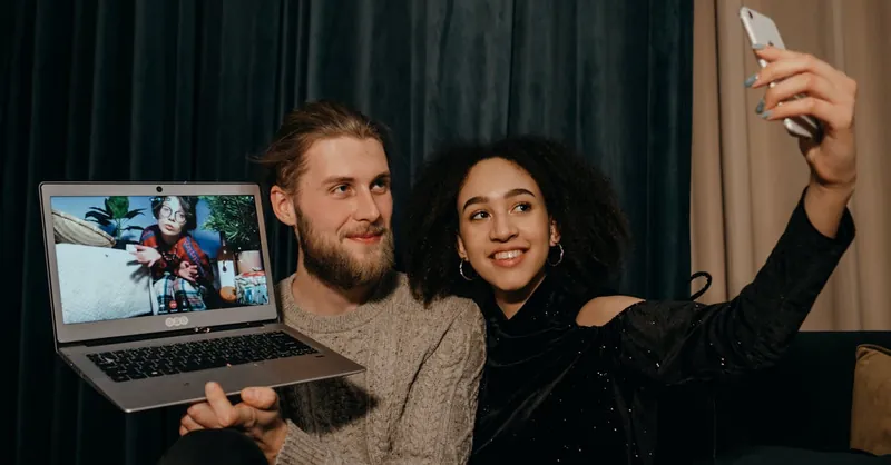A couple enjoying a video call while taking a selfie indoors with a laptop and smartphone.