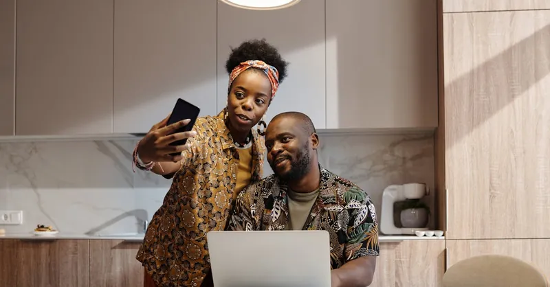 A cheerful couple making a video call in their kitchen using a smartphone and laptop.