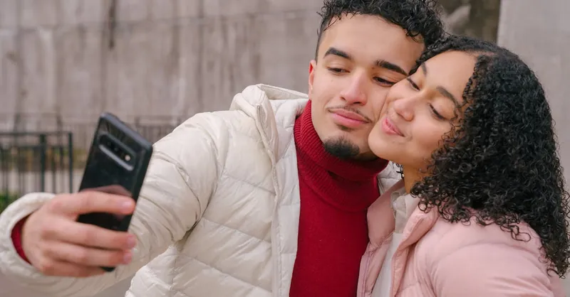 Enamored young Hispanic couple in warm casual clothes smiling and touching cheeks while taking selfie on mobile phone standing on street