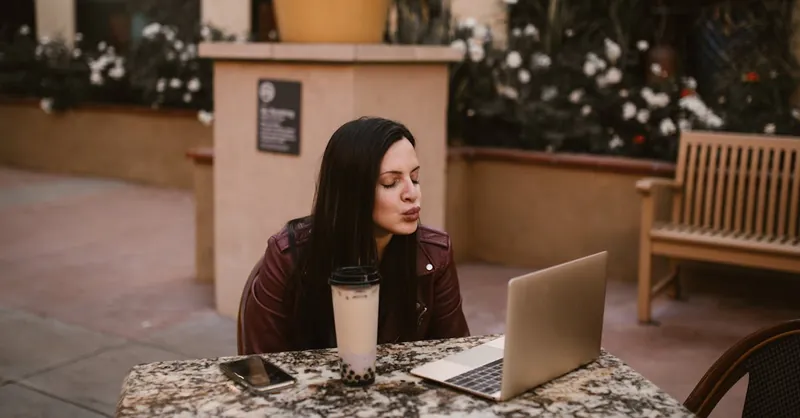 Young woman blowing a kiss during a virtual chat at an outdoor cafe table with laptop and bubble tea.