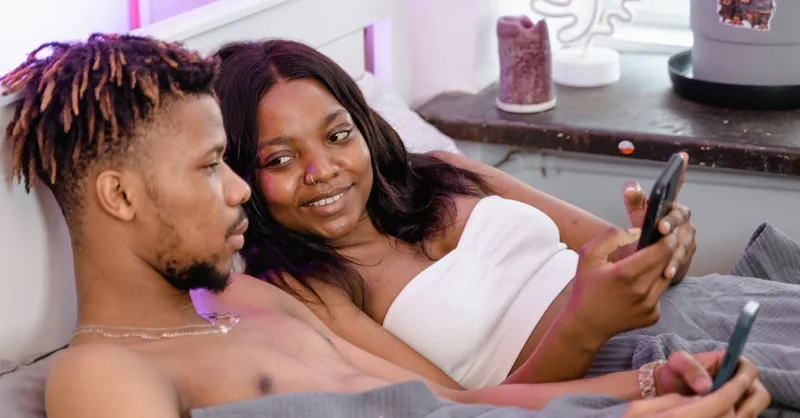 Young black couple lying in bed, using smartphones, showcasing modern intimacy and connection.