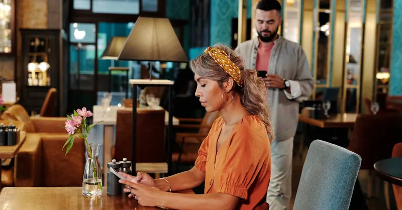 A couple on a first date at a modern café. The woman awaits, using her smartphone.