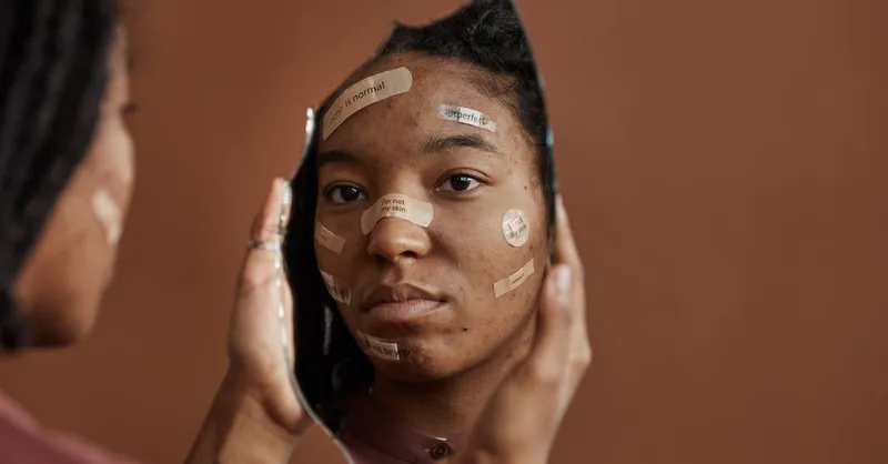 African American woman with Band-Aids looks into a shattered mirror, reflecting self-acceptance.