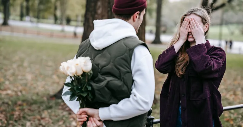 Young couple enjoying a romantic surprise moment in a sunny park, with hidden flowers and a vibrant autumn backdrop.